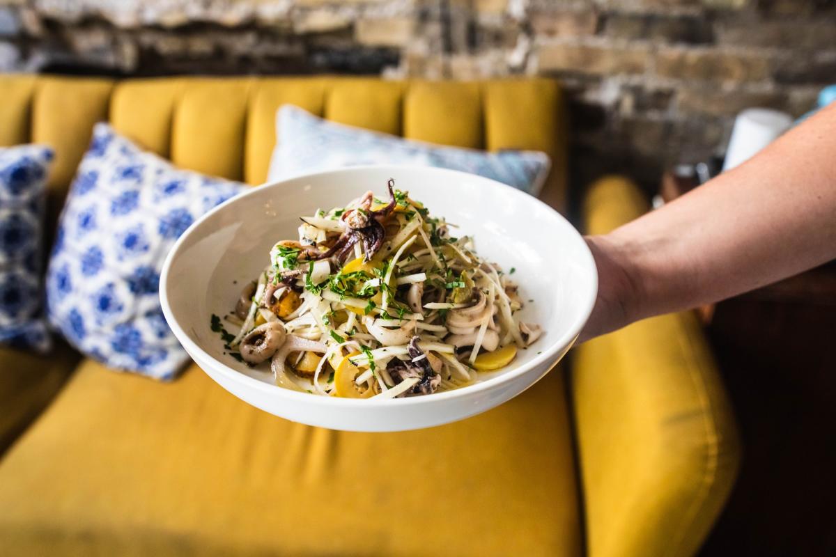 A person holds a white bowl filled with a seafood and vegetable dish, including squid, mushrooms, herbs and thinly sliced vegetables, at Amilinda in Milwaukee.