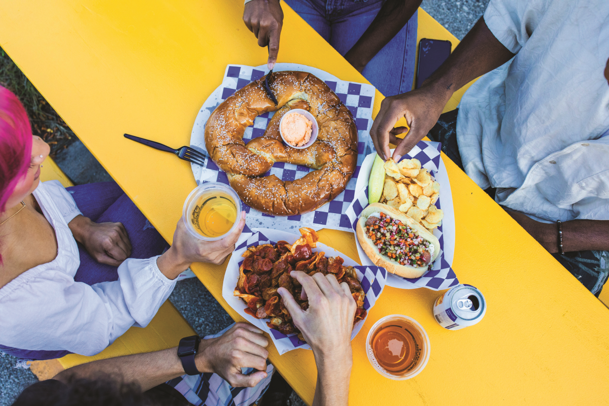 Friends share a giant soft pretzel, loaded fries, a hot dog, chips and drinks around a bright yellow outdoor table.