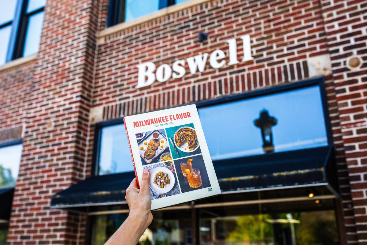 A hand holds up a copy of Milwaukee Flavor: The Cookbook in front of Boswell Book Company, a brick building with the store’s name in large white letters above the entrance.