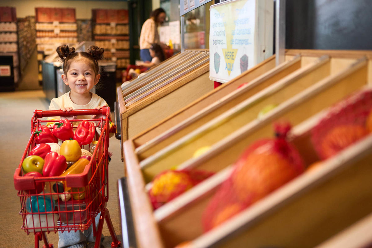 A young child smiles while pushing a red toy shopping cart filled with play fruits and vegetables at a grocery-themed exhibit in the Betty Brinn Children’s Museum.
