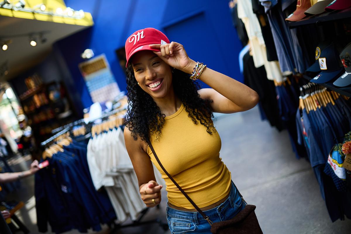 Smiling shopper trying on a red Miller-branded baseball cap inside the Miller Brewery gift shop, surrounded by racks of apparel and branded merchandise.