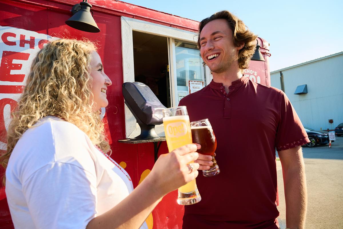 Two friends smile while holding pints of beer outside a red food truck, enjoying drinks together on a sunny afternoon at a brewery.