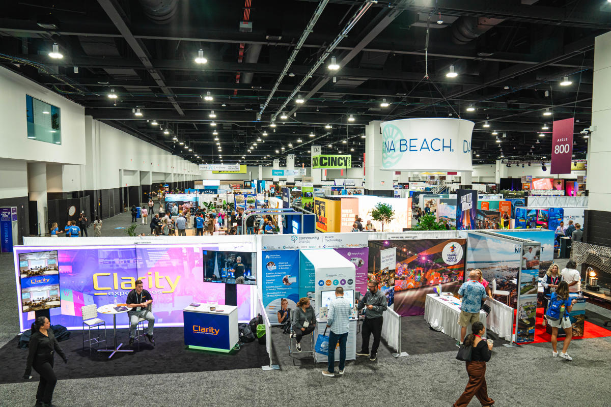 Wide overhead view of a busy convention tradeshow floor inside a large exhibition hall. Numerous exhibitor booths and destination displays fill the space, with attendees walking the aisles and talking with vendors. Large hanging signs mark different areas, including “Aisle 800,” and a prominent circular banner for a destination booth is visible above the crowd.
