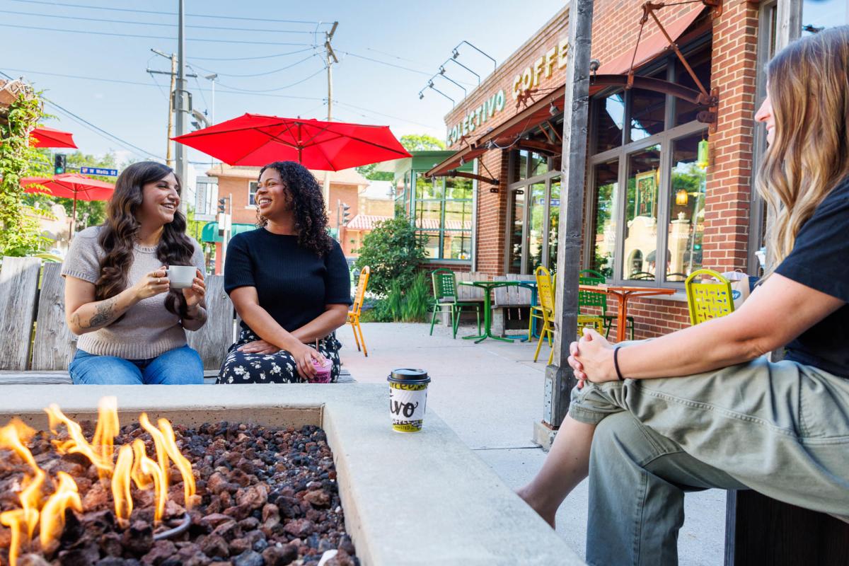 Three women chat over coffee around an outdoor fire pit at Colectivo Coffee.