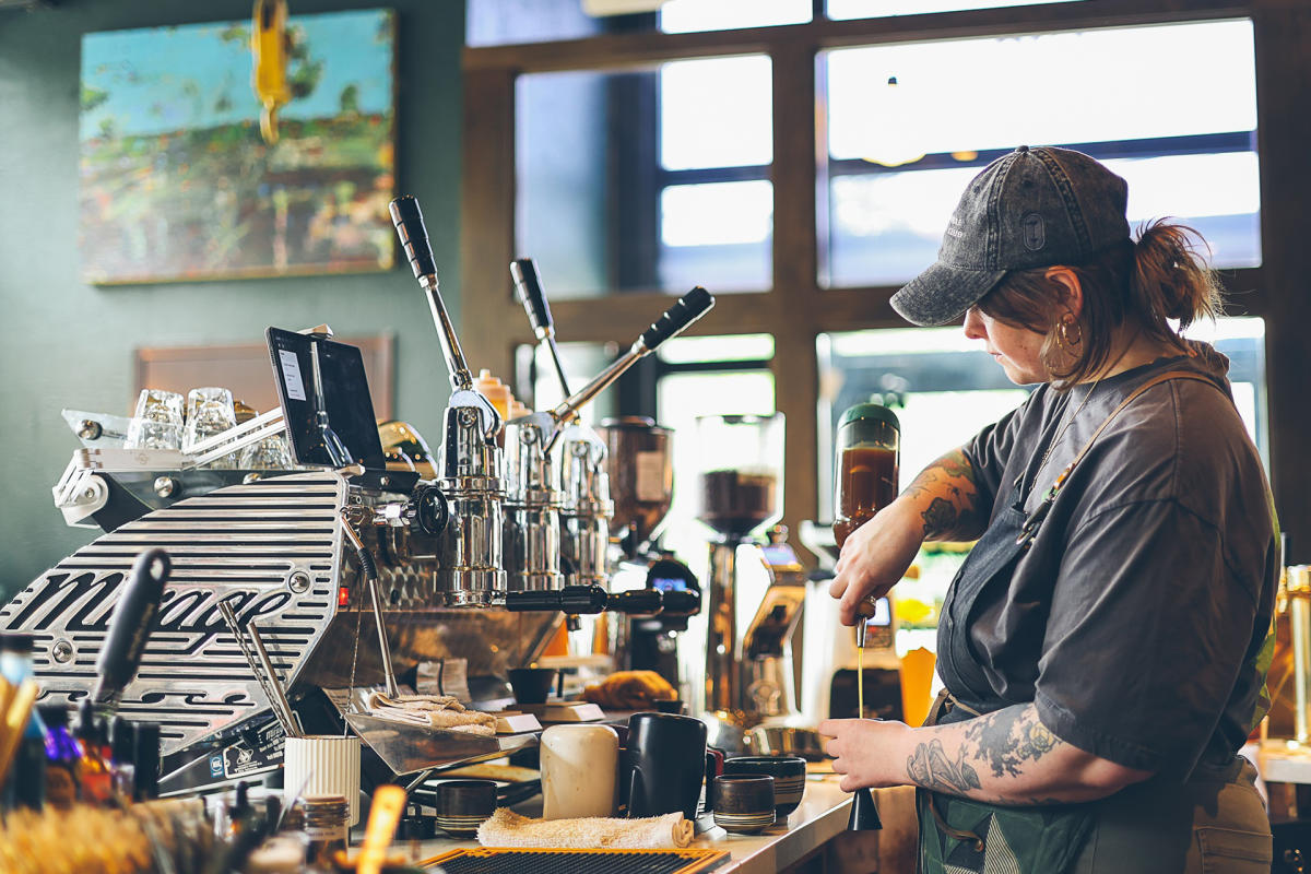 Barista preparing a drink at a coffee machine inside a café, surrounded by coffee-making equipment and supplies.