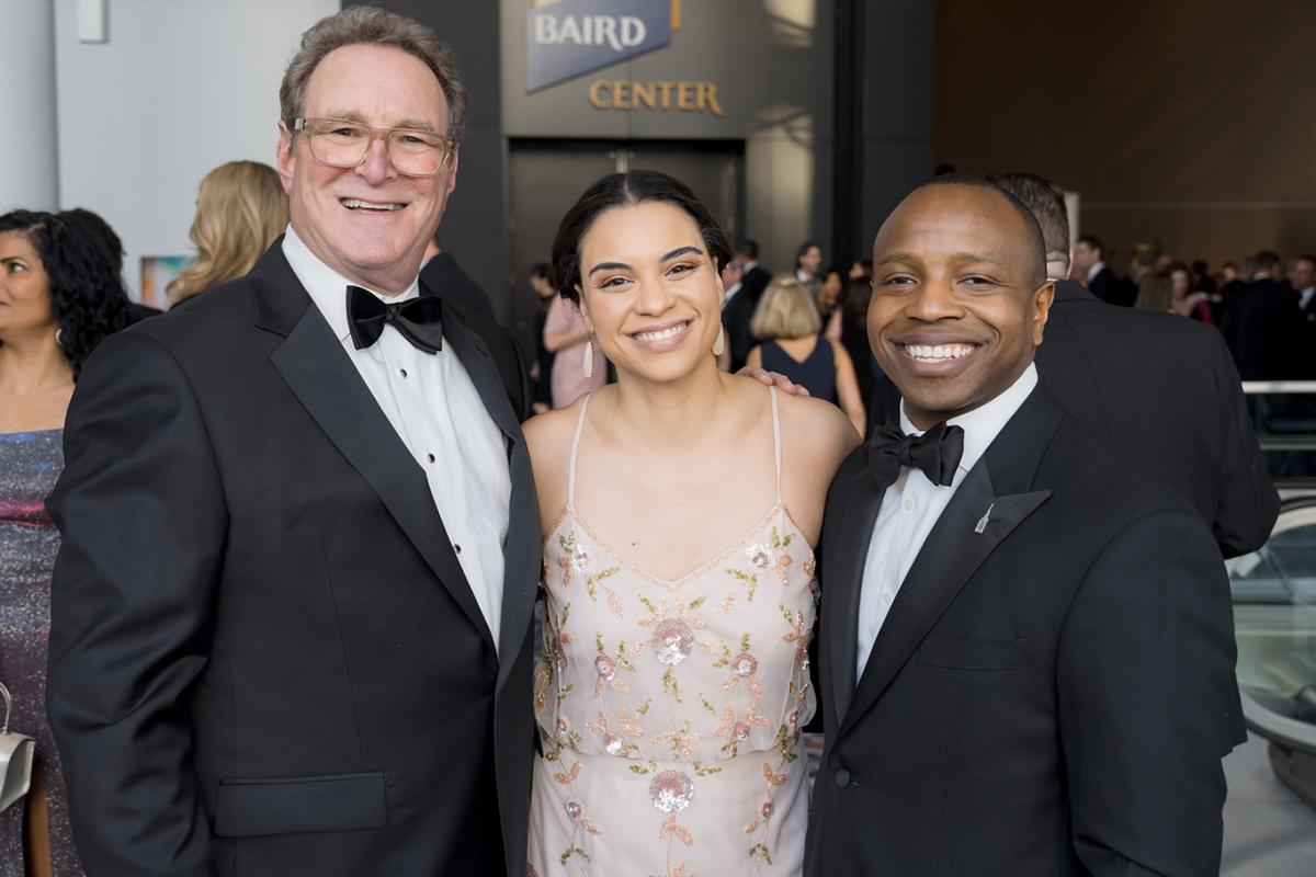 Photo of three people posing and smiling at a formal event inside the Baird Center. Two men in black tuxedos stand on either side of a woman in a light pink, floral embroidered dress. Other guests and event activity are visible in the background.