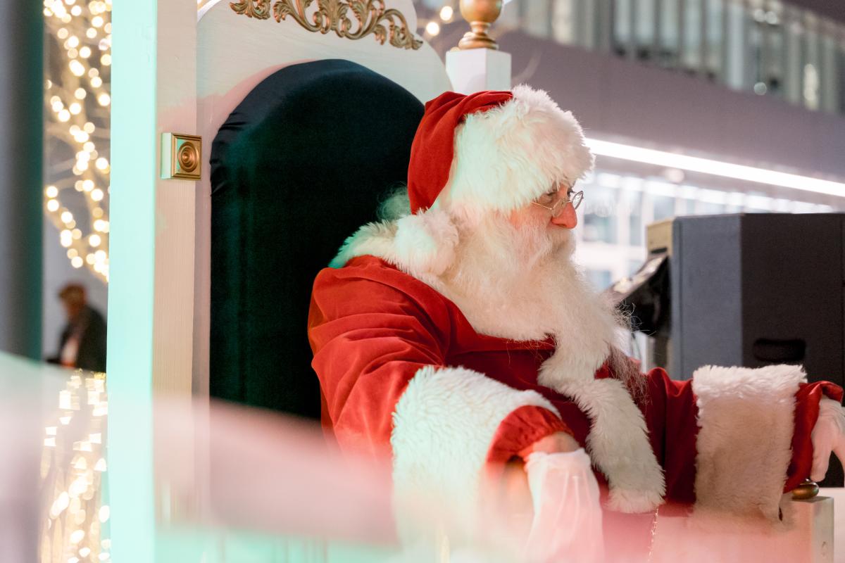 Santa Claus sits in a large, ornate chair wearing his traditional red suit and hat during an outdoor holiday event, with warm decorative lights glowing in the background.