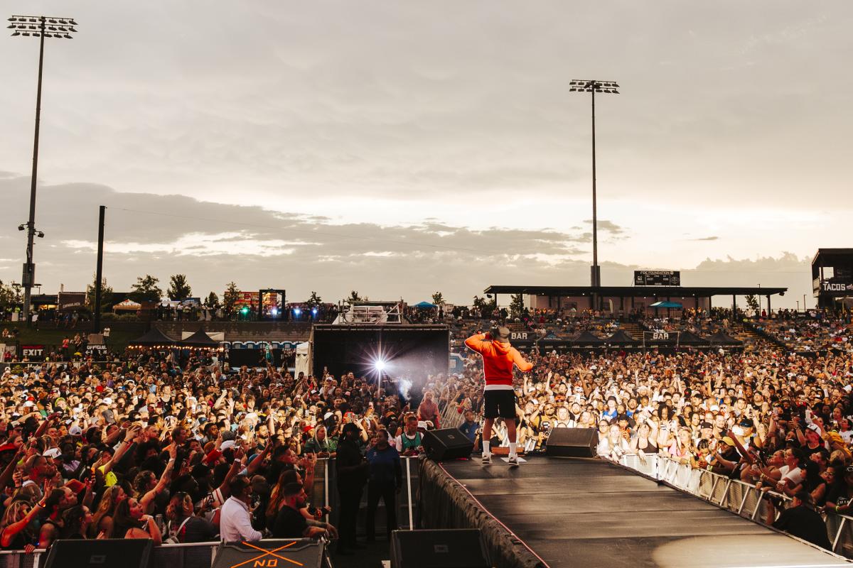 A performer in a red hoodie stands on an outdoor stage facing a massive crowd at sunset. Thousands of fans pack the venue, many holding up phones to capture the moment, while bright stadium lights and food stand signs glow in the background.