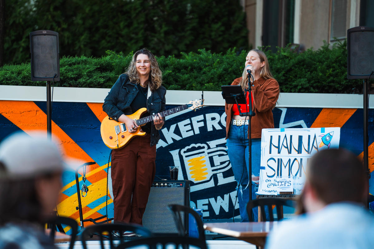 Two musicians perform outdoors in front of a colorful mural and a small audience. One person smiles while playing a yellow electric guitar, while the other sings into a microphone with eyes closed. Speakers flank the performance area, and a hand-painted tip sign reading "Hanna Simone" stands beside the singer. Blurred audience members and café-style tables are visible in the foreground.
