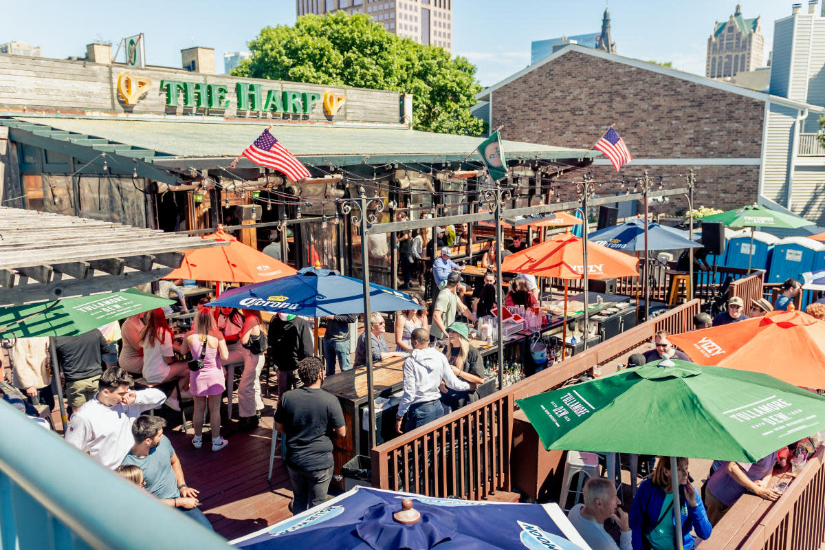 Outdoor patio at The Harp in downtown Milwaukee, filled with patrons seated beneath colorful umbrellas, with American flags and the city skyline in the background.