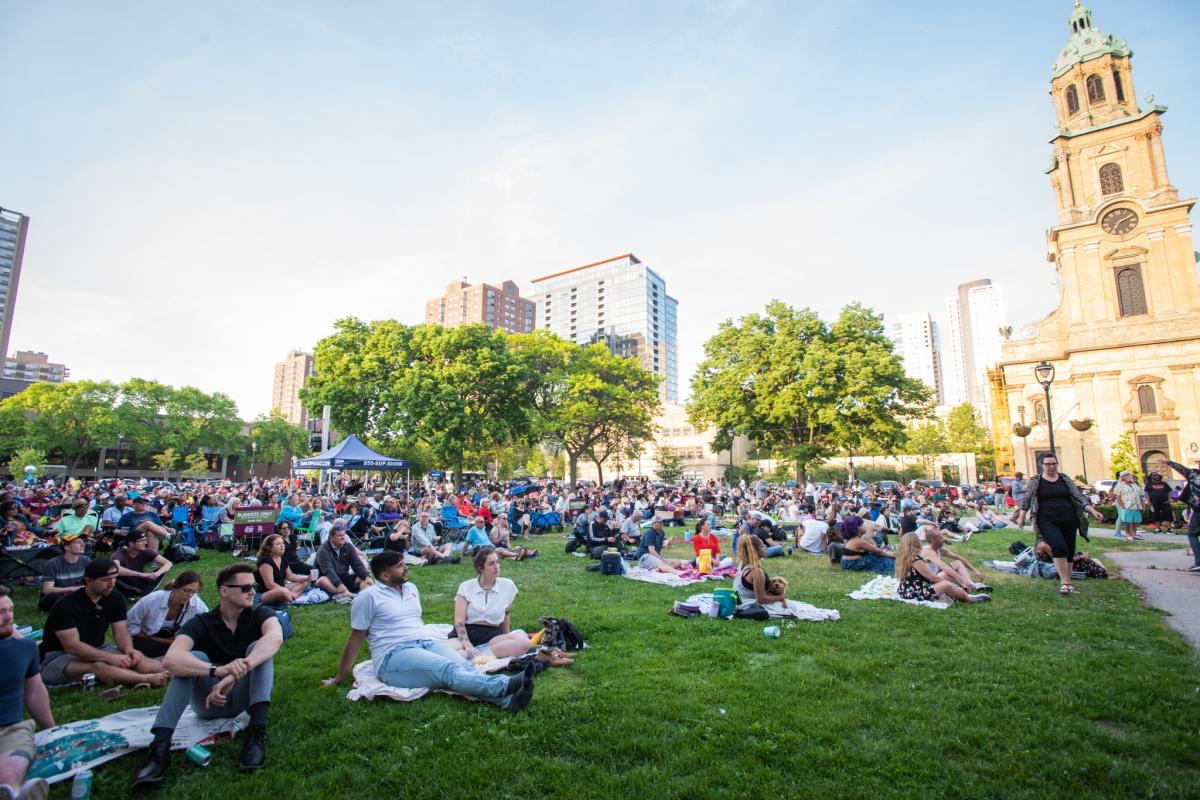A large crowd of people sitting on blankets and lawn chairs gathered on a grassy park area for Jazz in the Park, with a historic stone church with a green dome visible on the right side and modern city buildings in the background under a clear blue sky.