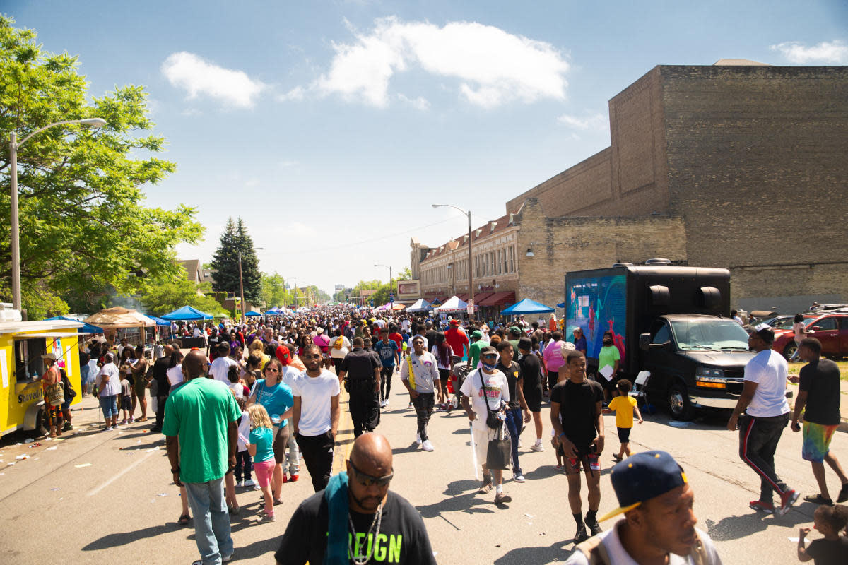 A large crowd fills a city street lined with vendor tents, food trucks, and historic buildings during a sunny outdoor Juneteenth celebration. People of all ages walk, talk, and enjoy the festive atmosphere under a blue sky with scattered clouds.