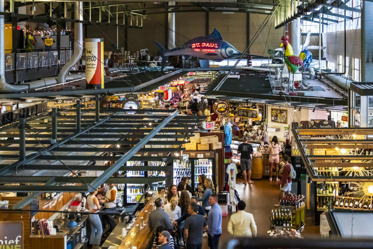 People stroll through vendors of the Milwaukee Public Market, seen from above with large illuminated sign it the background of the St. Paul Fish Company.