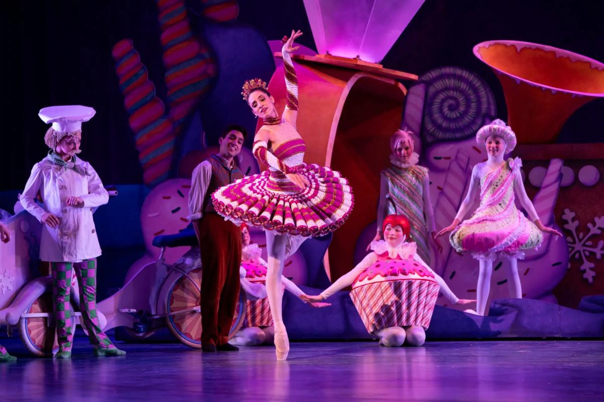 A ballerina in a red-and-white candy-themed costume performs a graceful pose on stage, surrounded by dancers dressed as sweets, including cupcakes and confectioners. The colorful set features oversized candies and whimsical decorations, illuminated by pink and purple stage lighting.