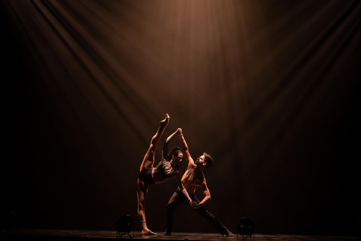 Two contemporary dancers perform an expressive lift under dramatic stage lighting.