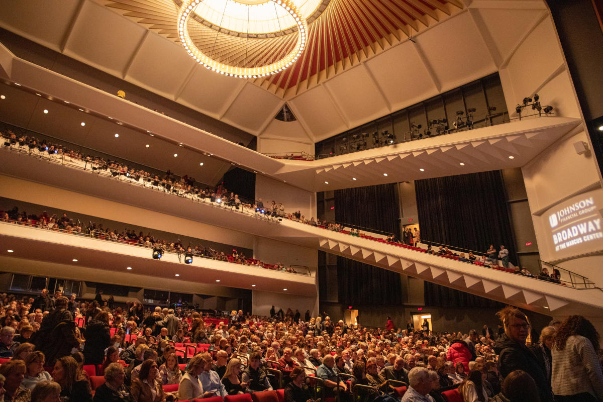 Interior of a crowded theater with multiple balcony levels, red seats, and a distinctive circular ceiling with ring chandelier