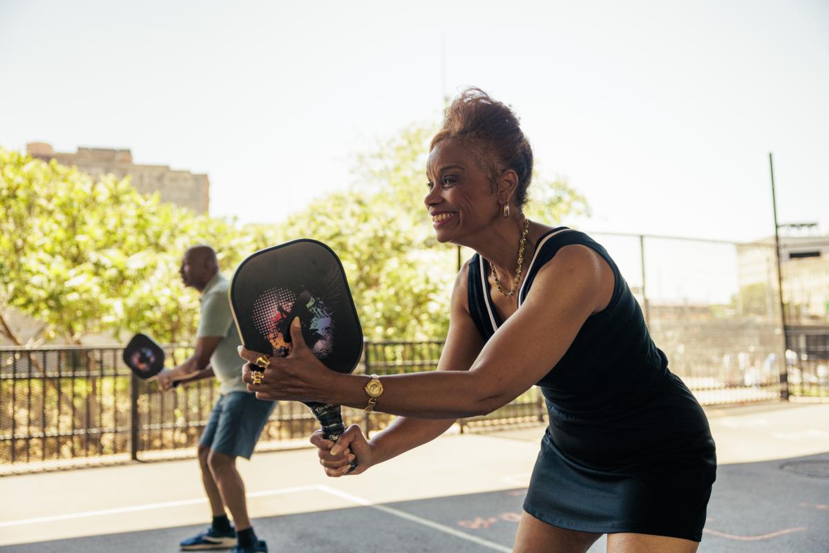 A smiling woman holds a pickleball paddle, ready to return a shot on an outdoor court, while another player stands behind her near a fence with trees and buildings in the background.
