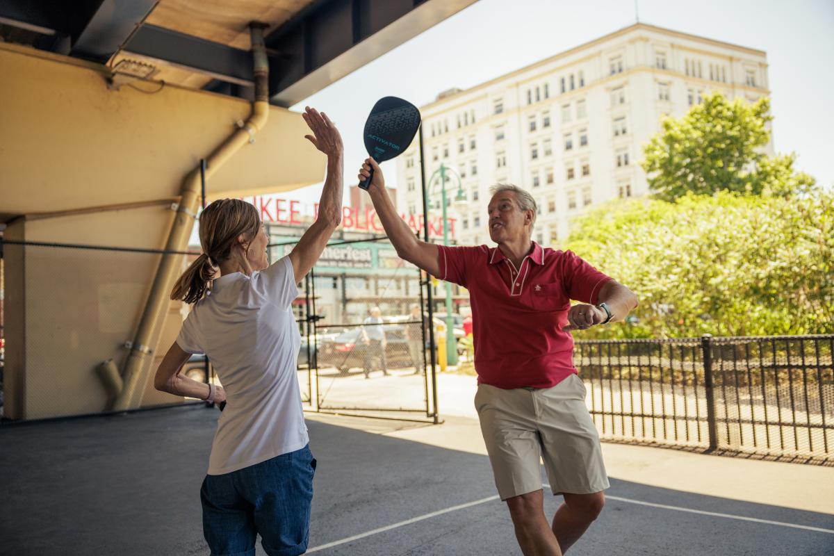 Two people celebrate with a high-five on an outdoor pickleball court beneath an overpass, with the Milwaukee Public Market sign and a city building visible in the background.