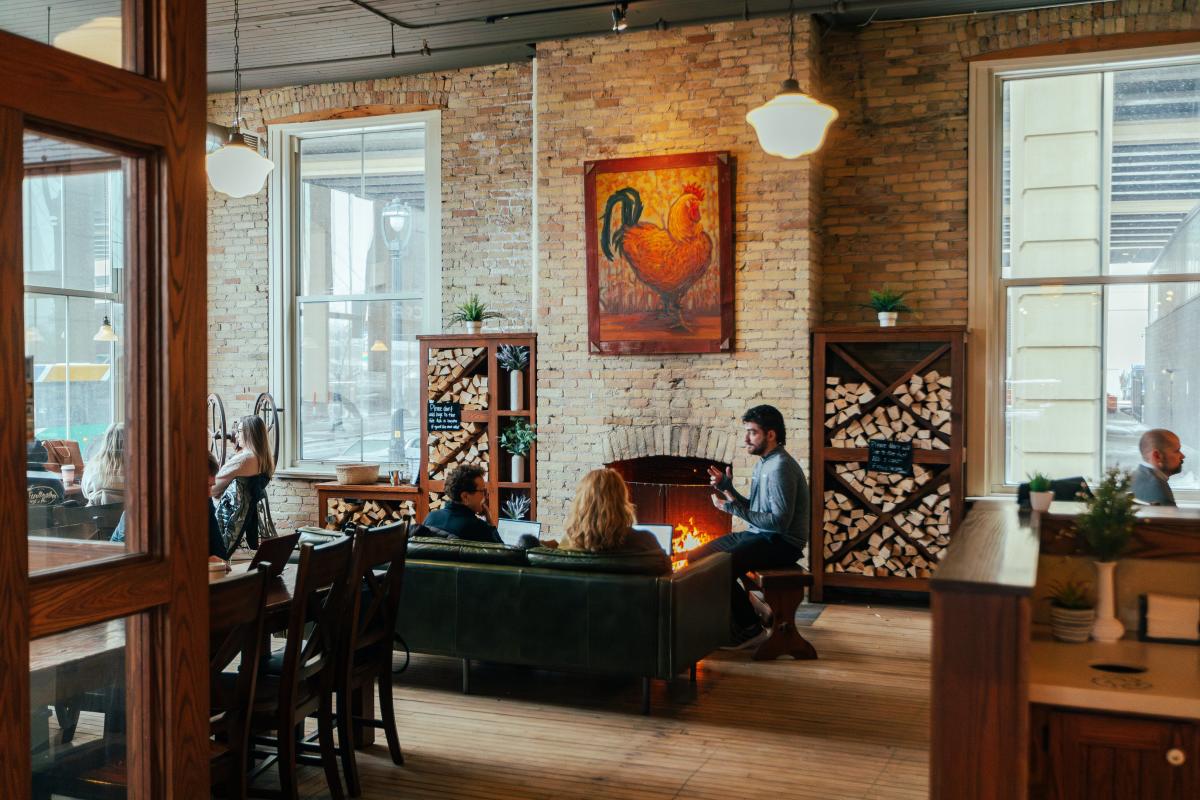 People sitting by a fireplace inside Stone Creek Coffee in Milwaukee, featuring brick walls, large windows, and cozy seating with a rooster painting above the mantel.
