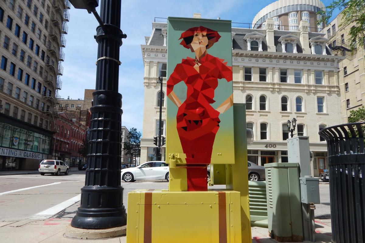 A utility box on a downtown Milwaukee street corner is painted with a bold geometric mural of a woman in a red dress and wide-brimmed hat, her hands on her hips in a confident pose. The faceted design uses angular shapes in red, orange, and brown, set against a gradient background of green and yellow. Historic buildings, cars, and a black lamppost surround the scene, capturing the blend of art and urban life.