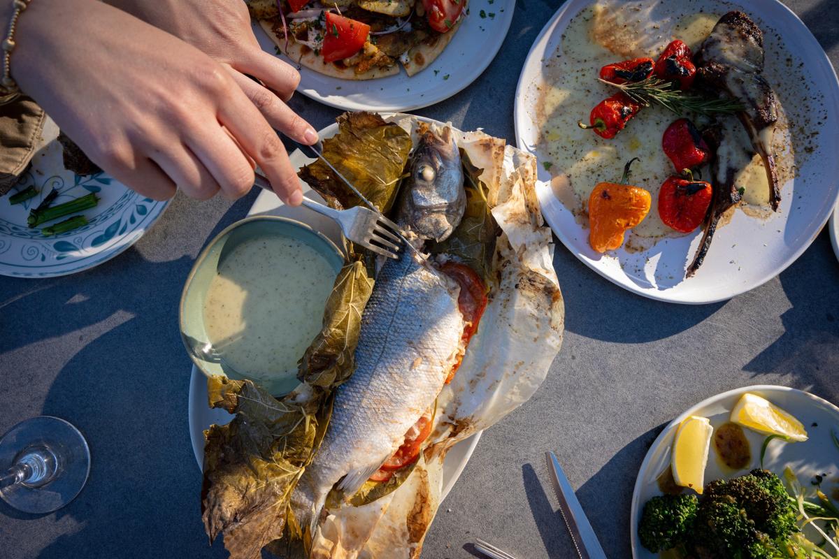 Overhead view of a restaurant table featuring several Mediterranean-style dishes. A person uses a fork and knife to cut into a whole baked fish wrapped in grape leaves and parchment paper, surrounded by plates of grilled vegetables, lamb chops with roasted peppers, broccoli, lemon wedges and a bowl of creamy sauce. Sunlight casts long shadows across the table.