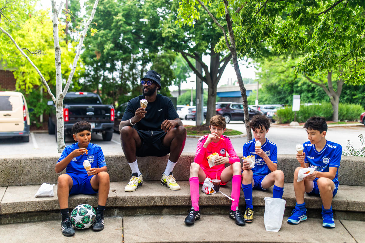 4 kids in soccer uniforms sit with Bobby Portis at Kopp's custard enjoying custard cones, french fries and chicken tenders. The background shows green trees and a parking lot.