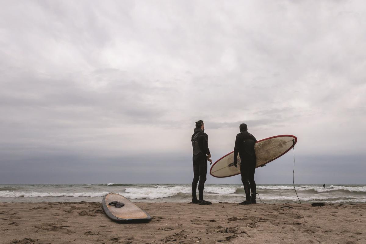 Two surfers in wetsuits stand on the beach looking toward Lake Michigan, with surfboards nearby under an overcast sky.