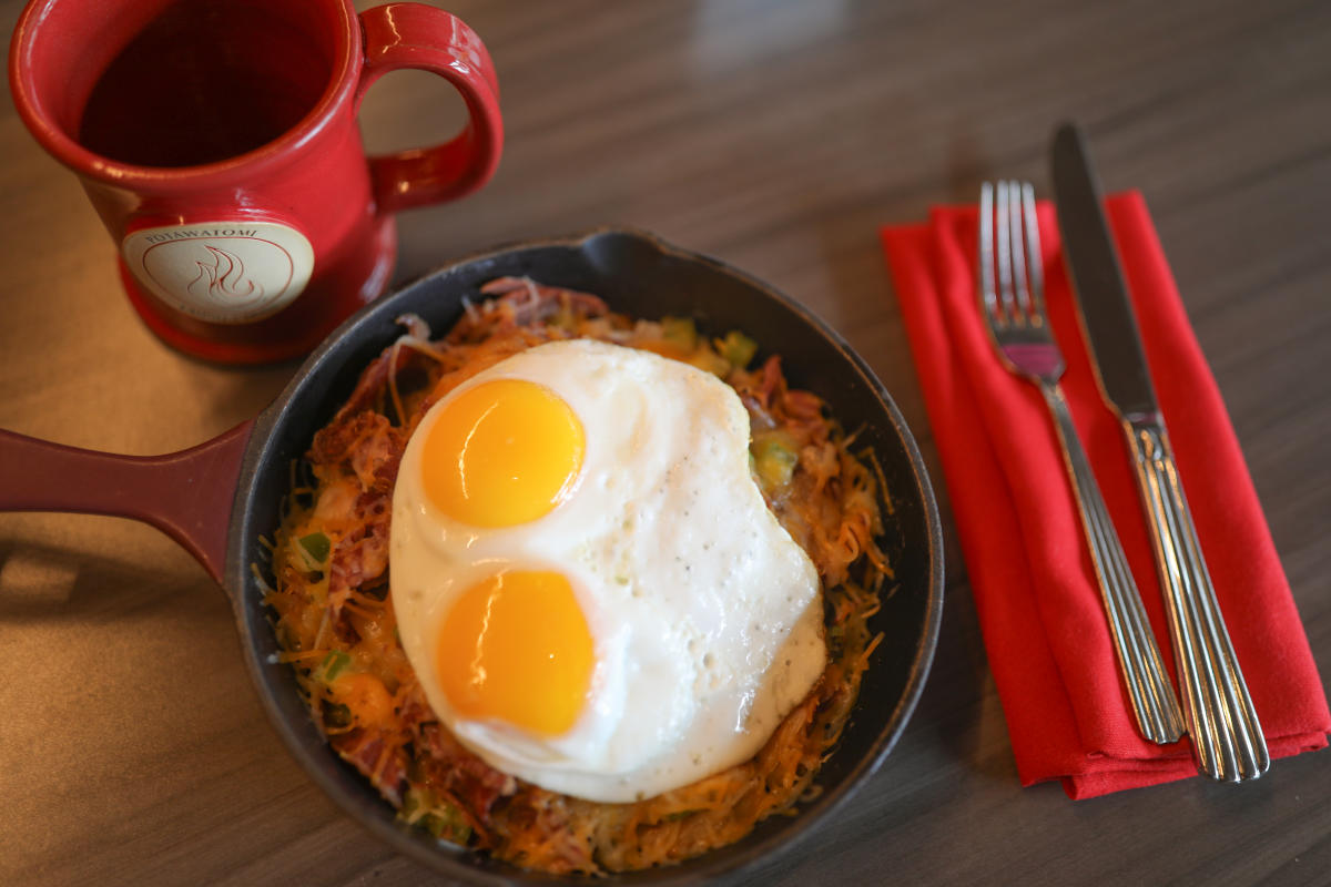 A skillet filled with corned beef hash topped with two sunny-side-up eggs, served with a red coffee mug and silverware wrapped in a red napkin on the side.