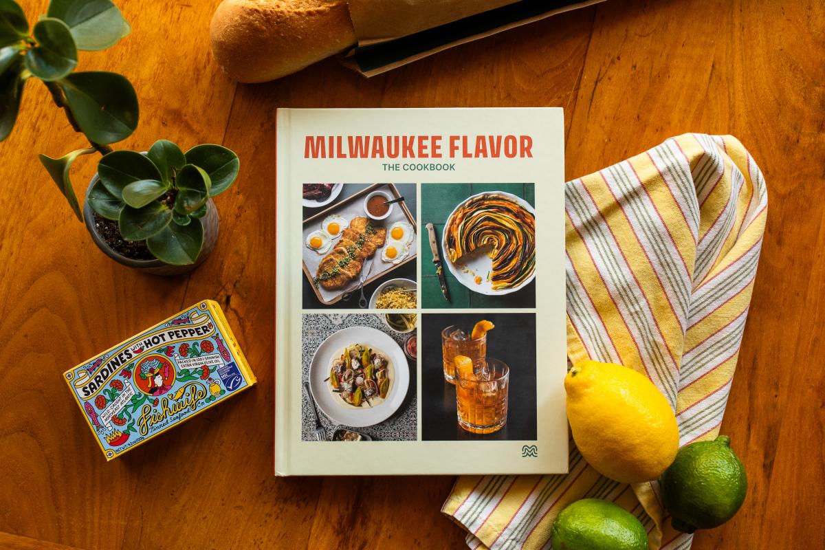 Milwaukee Flavor: The Cookbook displayed on a wooden table surrounded by colorful ingredients and props. A striped kitchen towel, a lemon, two limes, a loaf of bread, a potted plant, and a box of sardines with hot pepper frame the cookbook’s cover, which features photos of dishes and cocktails.