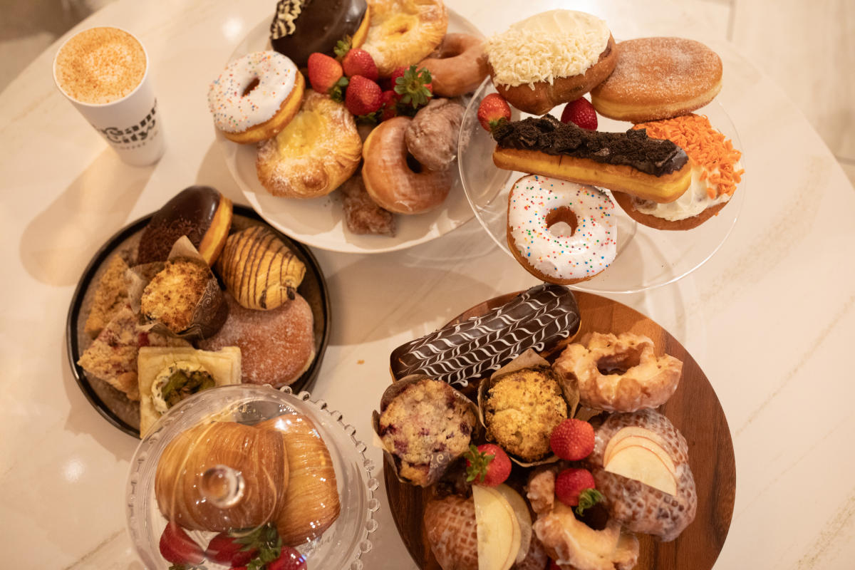 A table filled with assorted pastries and donuts on multiple trays and stands, including frosted donuts with sprinkles, eclairs, muffins, danishes, croissants, and fresh strawberries, with a cup of coffee on the side.