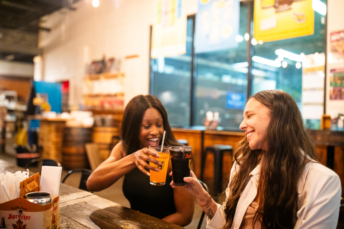Two women sitting at a wooden table in Sprecher Brewery, smiling and toasting with glasses of beer - one holding a light-colored beer and the other a dark beer. The setting appears warm and friendly with restaurant lighting and blurred background showing other seating areas and windows.
