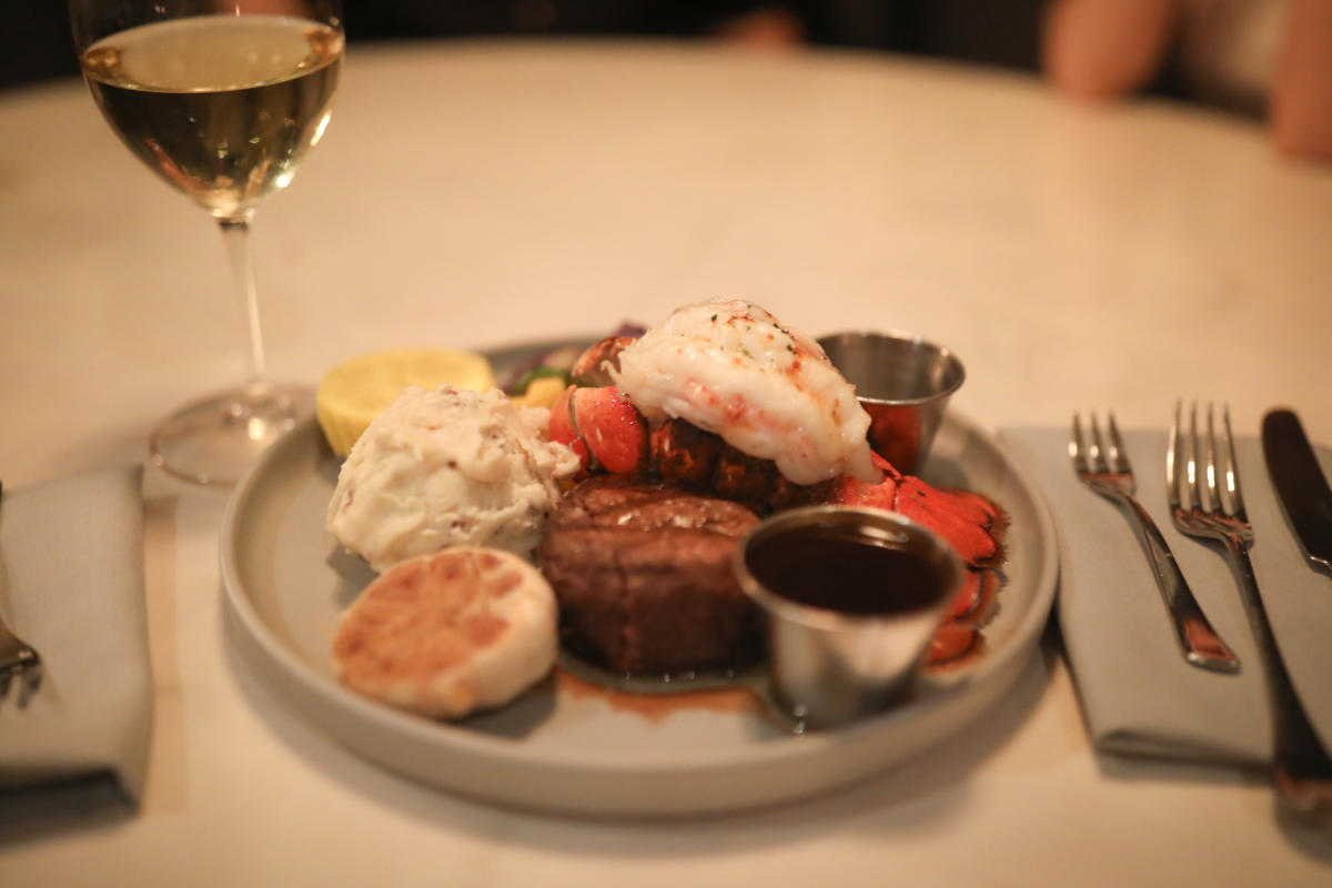 A surf and turf dinner plate with filet mignon, a lobster tail, mashed potatoes, roasted garlic, and dipping sauces, served with a glass of white wine on the side.