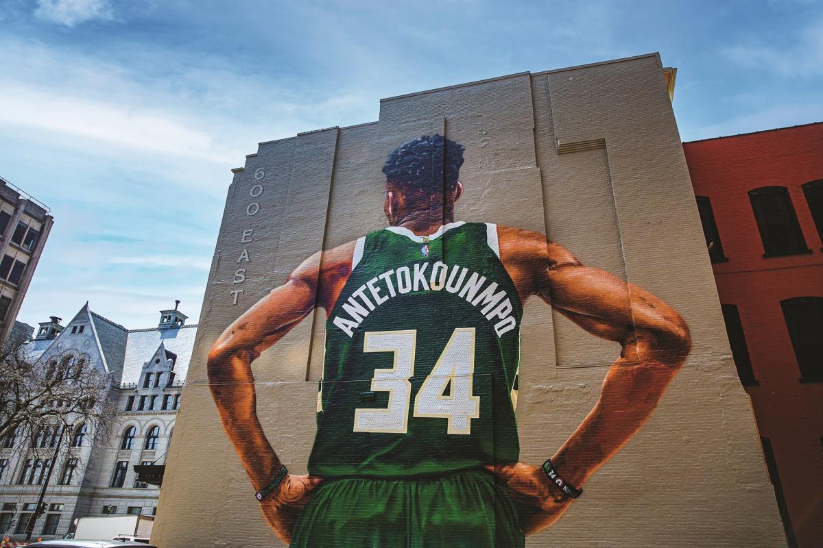 A large mural of Milwaukee Bucks player Giannis Antetokounmpo covers the side of a downtown building. Viewed from behind, he stands with his hands on his hips wearing a green No. 34 jersey with “ANTETOKOUNMPO” across the back. Historic buildings and a partly cloudy sky frame the scene.