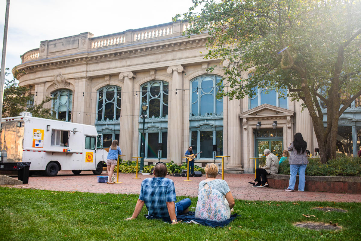 People relax and socialize on a lawn in front of a classical building with large arching windows, while a food truck is parked nearby. Sunlight filters through the trees, creating a warm atmosphere.