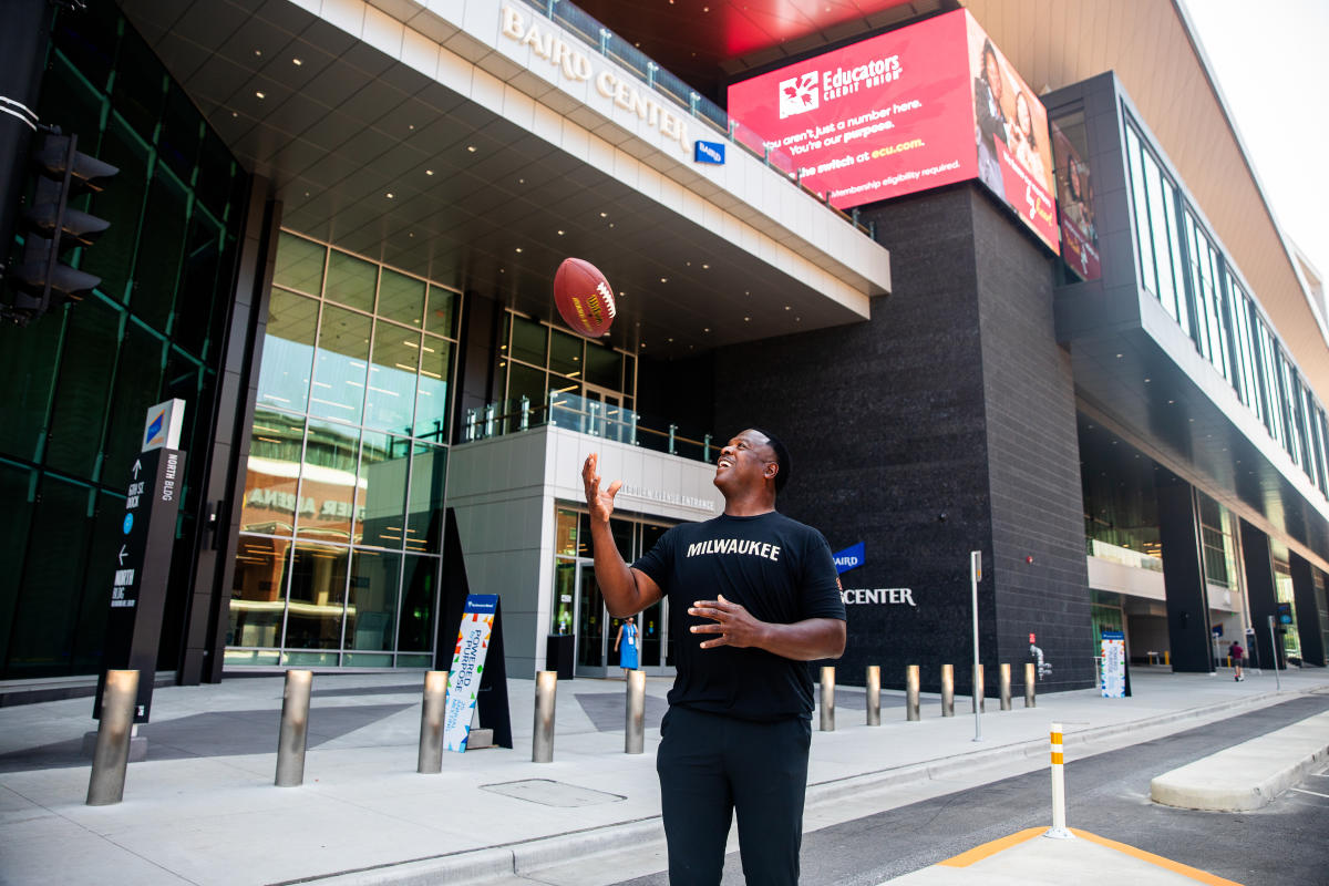 Outdoor photo of a man tossing a football in front of the Baird Center entrance. He is wearing a black shirt that says “Milwaukee” and smiling as the football is in midair above his hands. The modern convention center building, glass facade, and a large digital billboard are visible in the background.