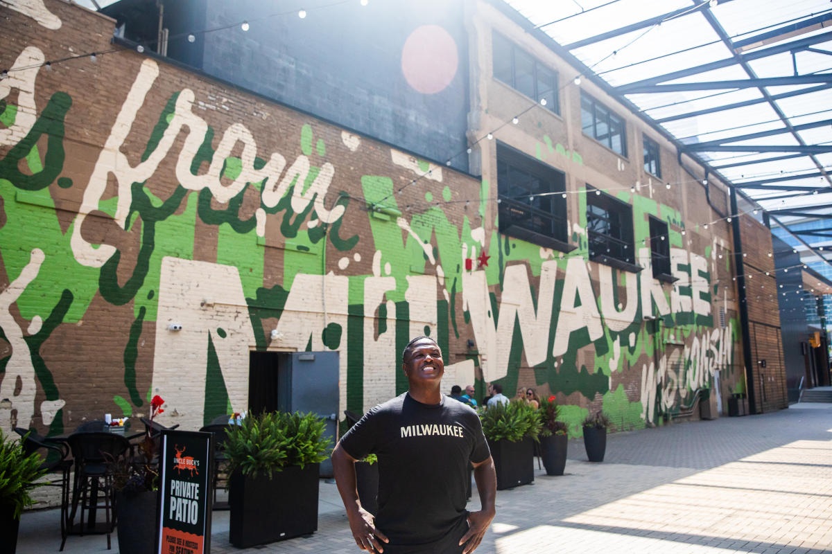 LeRoy Butler wearing a black Milwaukee T shirt stands smiling in a covered outdoor courtyard, with a large Milwaukee mural painted across the brick wall behind him and string lights overhead.