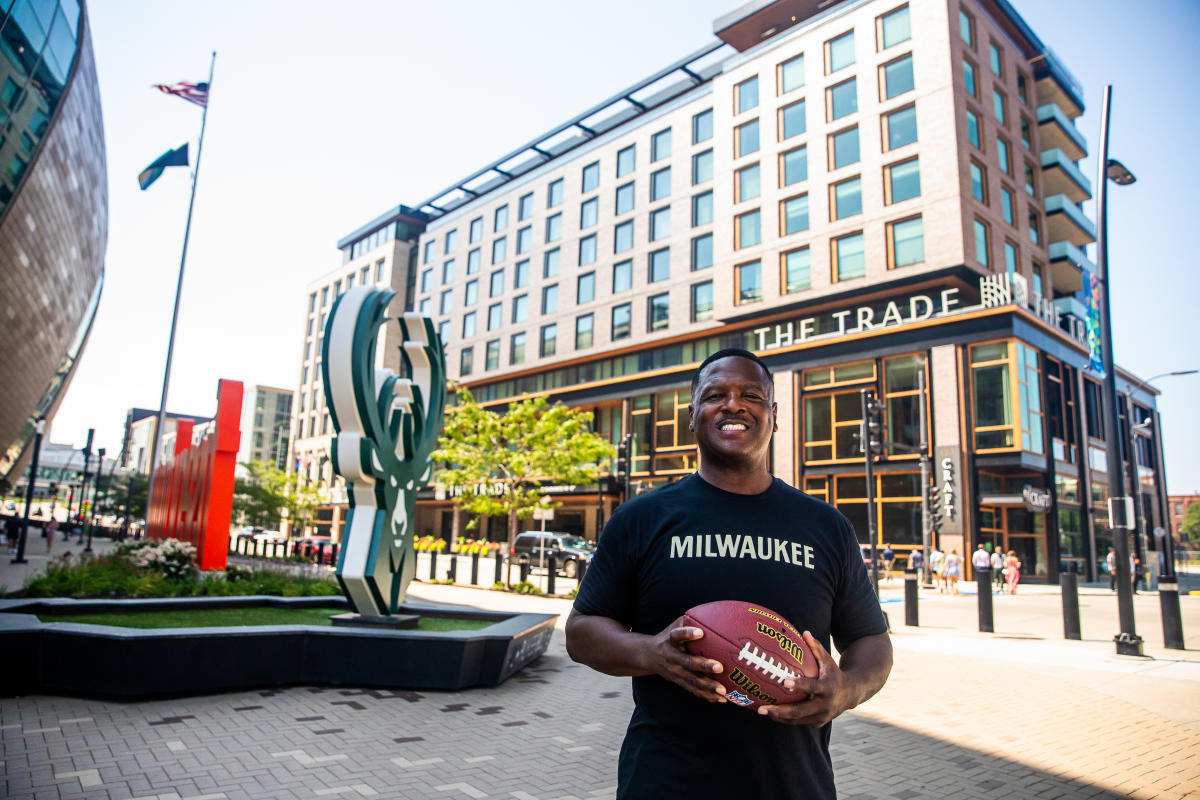 LeRoy Butler smiles while wearing a black ‘Milwaukee’ T-shirt holds a football in an outdoor plaza, with The Trade Hotel and a large deer sculpture visible behind him in Deer District.