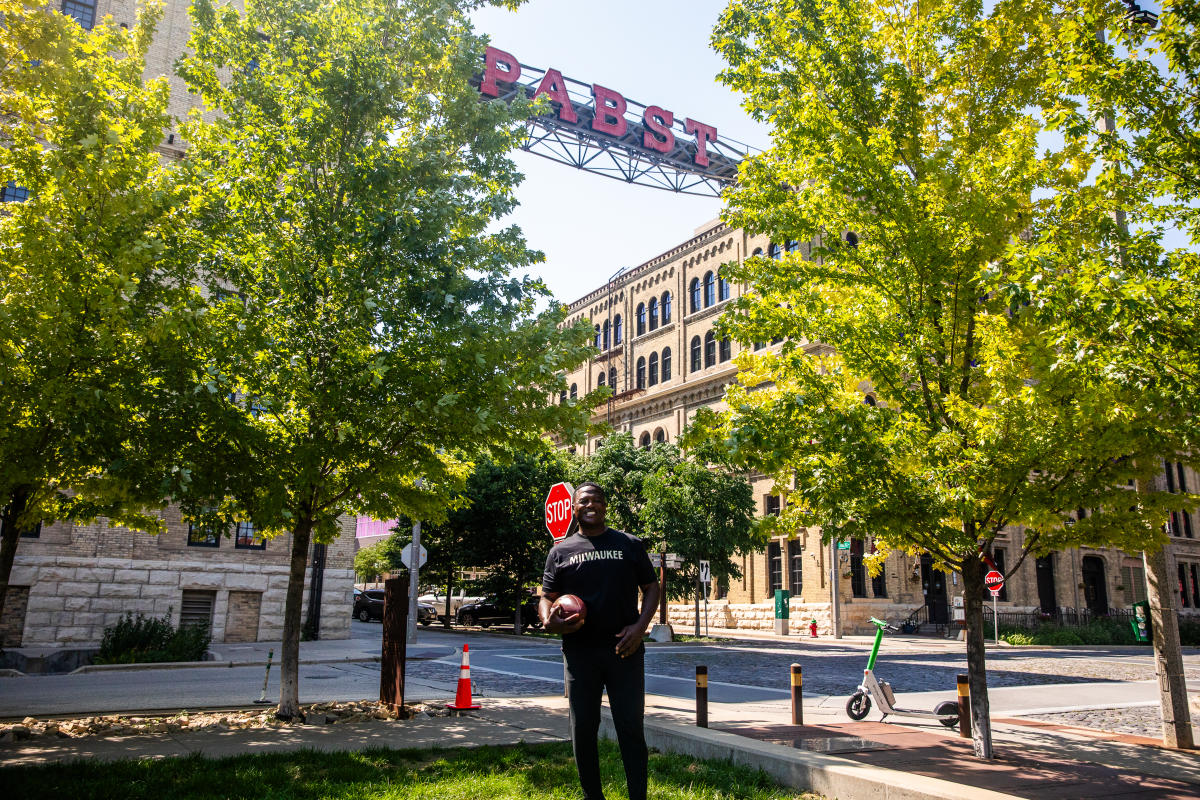 LeRoy Butler wearing a black Milwaukee T shirt stands holding a football beneath the Pabst sign, with historic brick buildings and leafy trees surrounding the street scene.