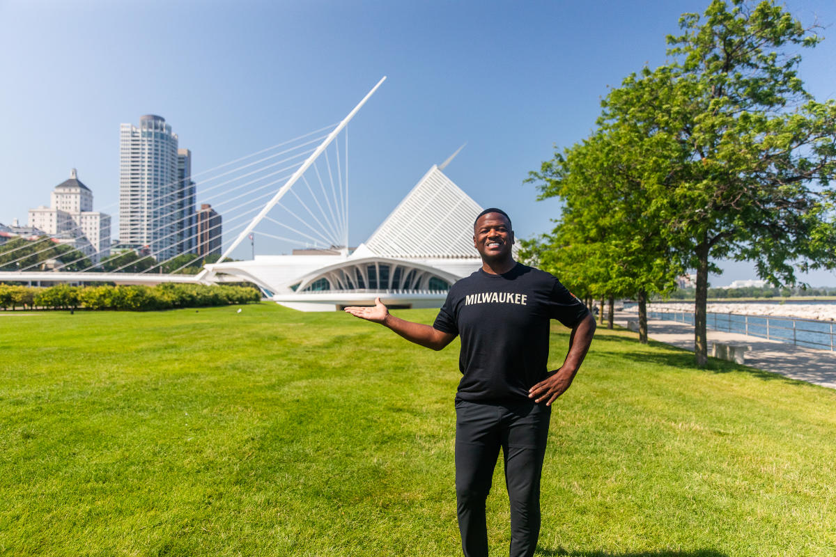 LeRoy Butler wearing a black Milwaukee T shirt stands on a grassy lakefront lawn, gesturing toward the Milwaukee Art Museum with the city skyline and Lake Michigan visible behind him.