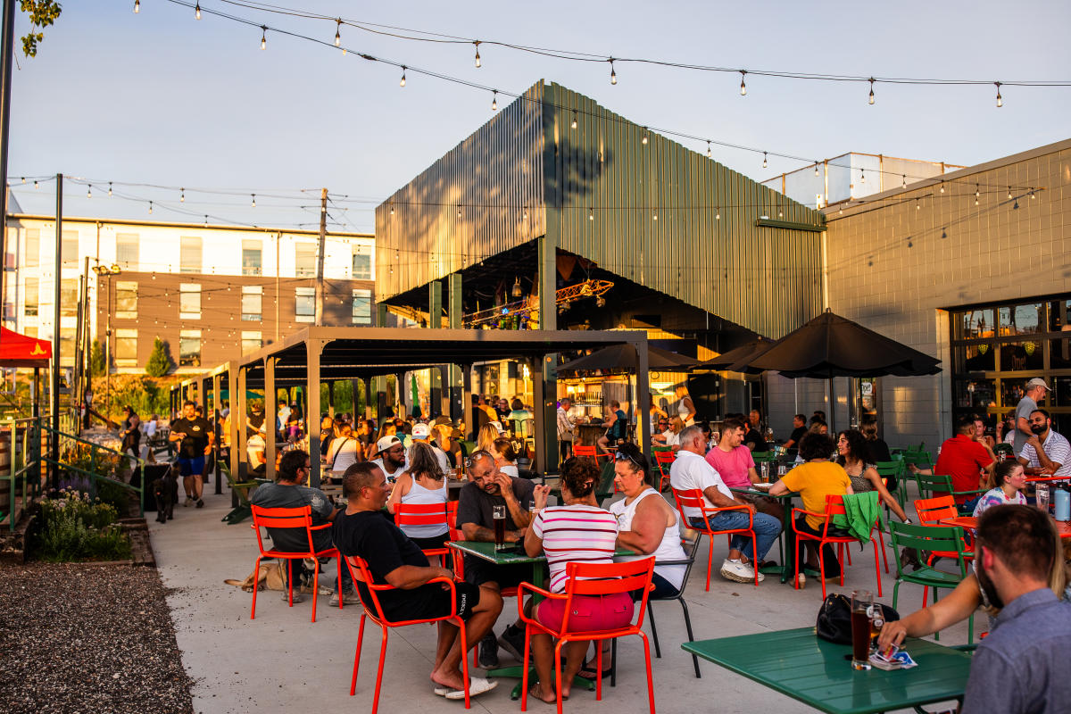 A large patio at a brewery is full of people sitting in red chairs. The golden light of sunset spills over the space. A large bar is in the background with a green overhang.