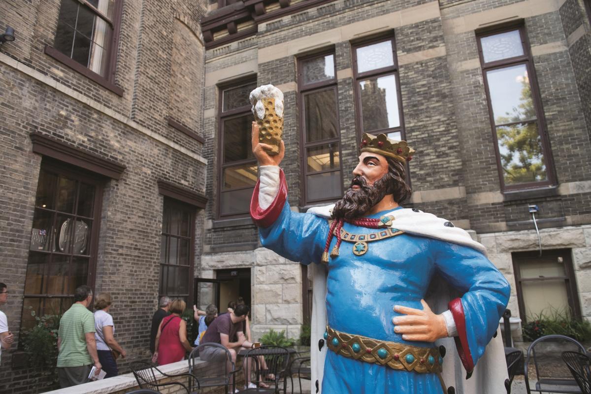 A colorful statue of King Gambrinus, the legendary beer king, holding up a frothy mug of beer in a celebratory pose outside the Historic Pabst Brewery in Milwaukee. People are gathered nearby in the brick courtyard.