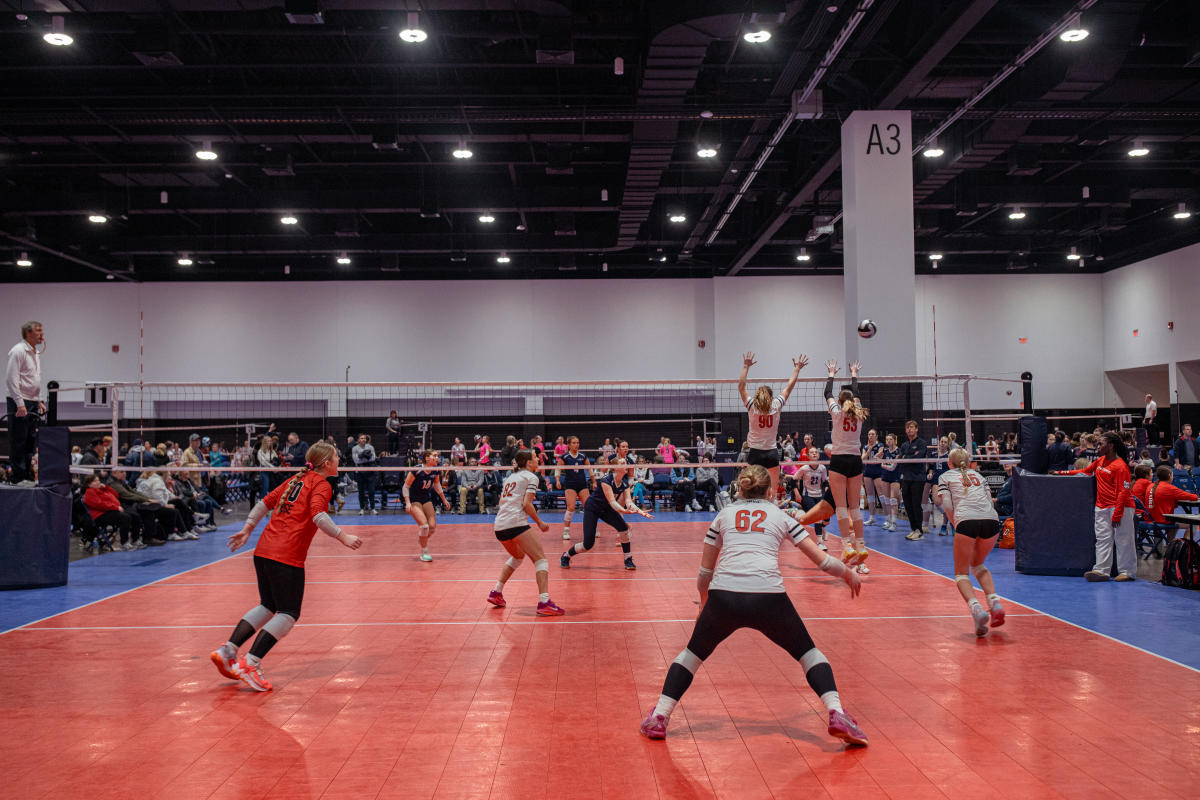 A competitive indoor volleyball match in progress at a large convention center court. Two players from the team in white jerseys leap to block the ball while their teammates prepare in defensive stances. The opposing team, wearing dark uniforms, readies to spike or return the ball. Spectators line the perimeter of the court, and the space is marked by bright lighting and high ceilings with an A3 column visible in the background.