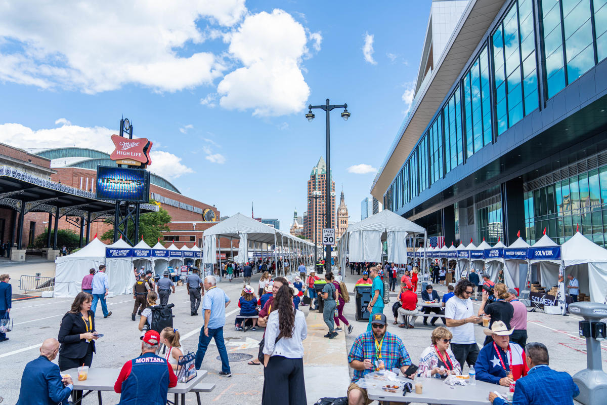 Wide street scene of an outdoor event in downtown Milwaukee. People walk and sit at tables between rows of white vendor tents set up along the street. The Baird Center’s glass exterior lines the right side of the image, and the Miller High Life Theatre sign and city skyline are visible in the background under a bright blue sky.