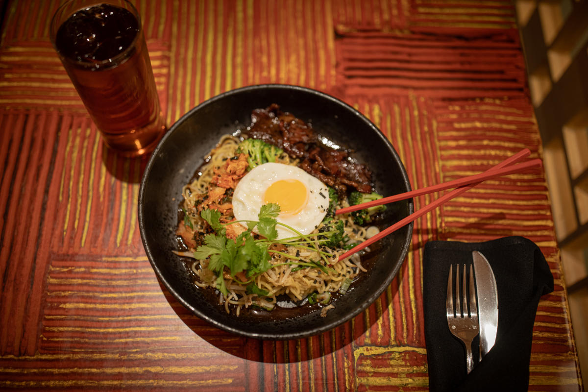 A bowl of noodles topped with a sunny-side-up egg, fresh cilantro, and crispy garnishes, served with slices of glazed meat and broccoli. Red chopsticks rest on the bowl, with a glass of iced tea and a wrapped fork and knife on the side, all set on a red and yellow patterned table.