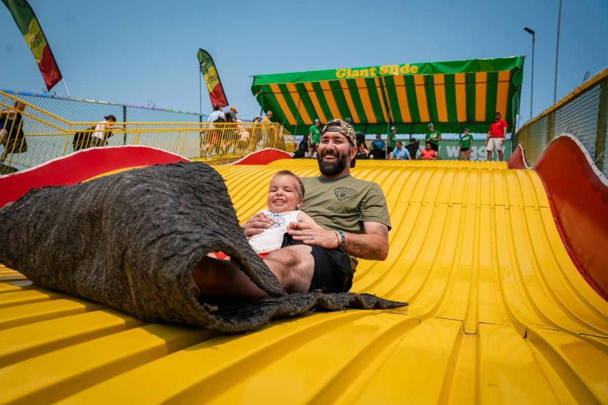 A smiling man holds a young child as they ride down the bright yellow Giant Slide together on a burlap mat at the Wisconsin State Fair.