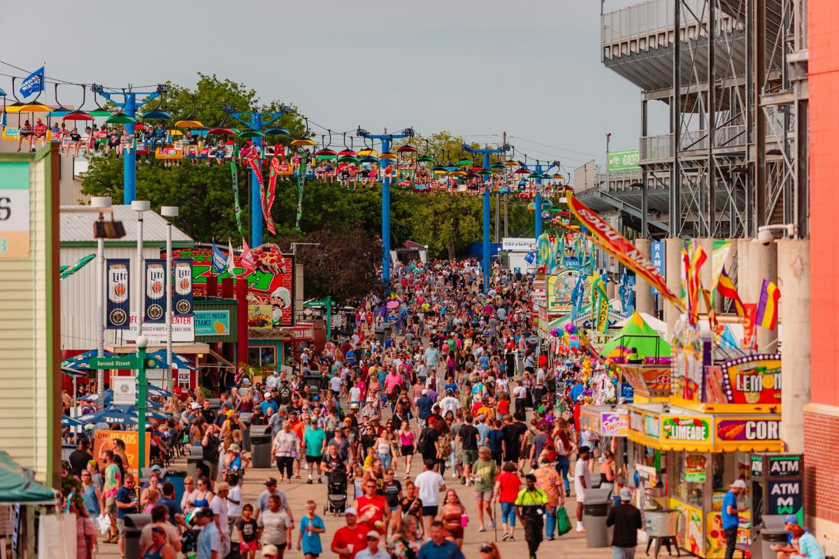 A dense crowd walks along Grandstand Avenue between food stands and vendor booths while riders pass overhead on the Sky Glider at the Wisconsin State Fair.