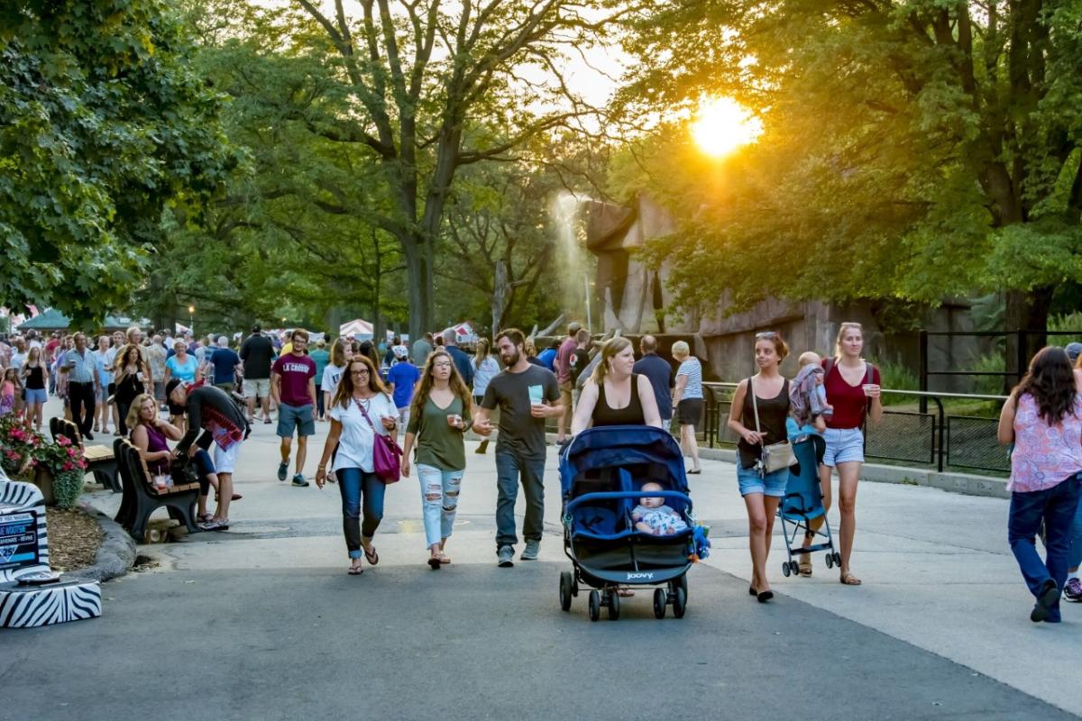 Visitors stroll through the Milwaukee County Zoo during a summer evening event, with the sun setting behind trees and a fountain in the background.