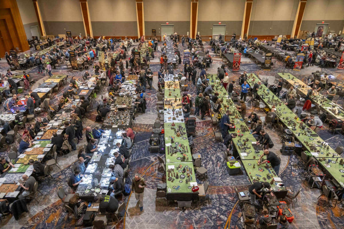 A large tabletop gaming convention in a spacious event hall, filled with rows of long tables where attendees play strategy and miniature-based games. The room buzzes with activity as participants move between tables covered in detailed game boards, terrain, and figurines. Colorful banners and displays surround the space, while the patterned carpet and neutral walls frame the energetic, community-driven environment.
