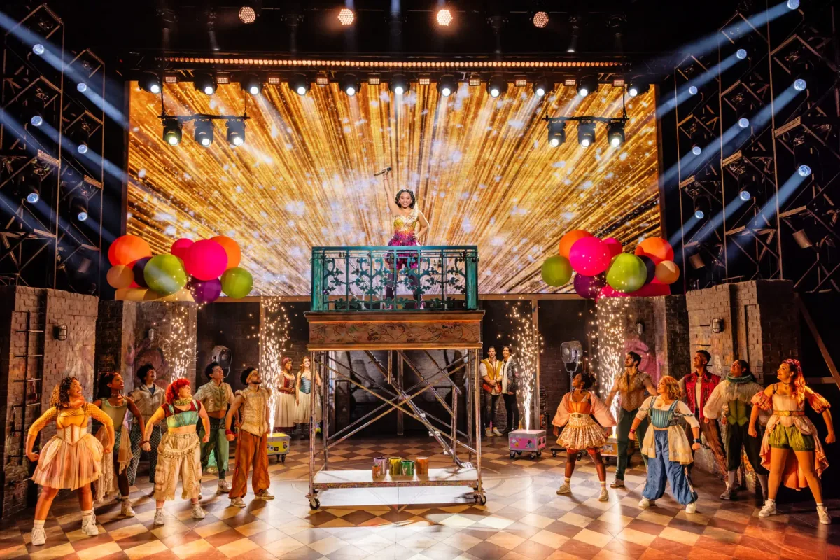 A large ensemble performs onstage at the Marcus Performing Arts Center during a colorful musical number. One performer stands atop a raised balcony platform at center stage holding a microphone, framed by a gold starburst LED backdrop and bright balloon clusters. Sparks shoot upward on both sides of the platform while dancers in vibrant costumes pose across the checkered stage beneath sweeping spotlights.