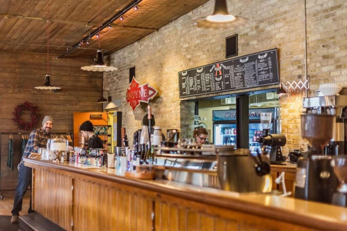 Interior of a cozy coffee shop with a long wooden counter, baristas preparing drinks, and a large chalkboard menu on a brick wall.